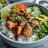 Colorful Baked Salmon Rice Bowl topped with edamame, green onions, and sesame seeds, drizzled with spicy sriracha mayo and fresh lime wedges.