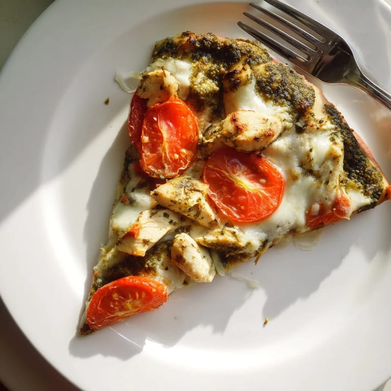 A close-up of a delicious Chicken Pesto Pizza with a crispy edge, showcasing shredded chicken, fresh basil leaves, and colorful tomato pieces.